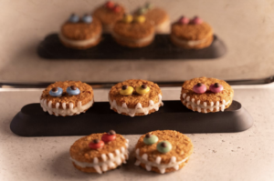 Detailne foto vegan Halloweeni koletisküpsistest, mis on kaunistatud värvilise glasuuri, nööpide ja šokolaadilaastudega, asetatud puidust lauale.