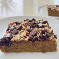 Gluteenivaba kõrvitsa-õuna piparkoogikook šokolaadinööpide ja pähklitega valgel taldrikul.